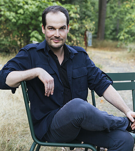 Portraitfoto von Max Czollek. Er sitzt seitlich auf einer dunkelgrünen Bank. Er hat dunkles Haar und einen Dreitagebart. Er trägt ein dunkelblaues Hemd über einem schwarzen T-Shirt. Hinter ihm erstreckt sich eine Wiese.