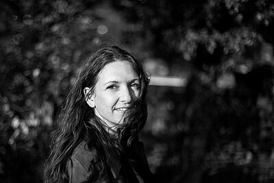 Schwarzweißes Portraitfoto von Carola Volles. Sie steht seitlich und blickt in die Kamera. Ihr welliges Haar fliegt ihr ins Gesicht. Der Hintergrund ist verschwommen, es sind Büsche wahrzunehmen.