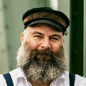 Portraitfoto von Ulrich Burdack. Er trägt ein weißes Hemd, dunkle Hosenträger, eine Schiffermütze sowie einen grauen Vollbart. Seine Arme sind vor der Brust verschränkt.