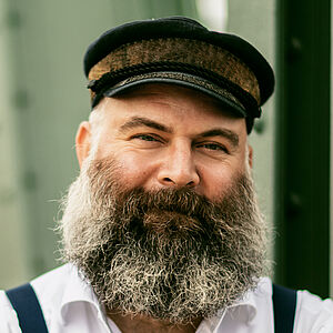 Portraitfoto von Ulrich Burdack. Er trägt ein weißes Hemd, dunkle Hosenträger, eine Schiffermütze sowie einen grauen Vollbart. Seine Arme sind vor der Brust verschränkt.