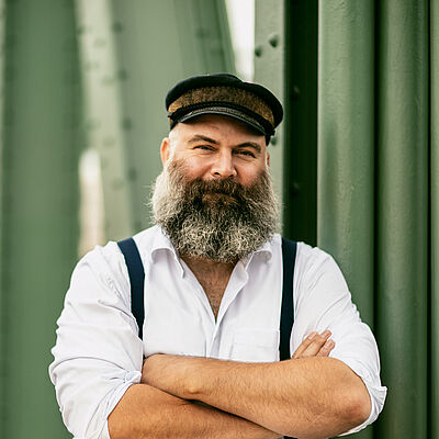 Portraitfoto von Ulrich Burdack. Er hat die Arme verschränkt. Er trägt einen Kapitänsmütze, einen grauen Vollbart, ein weißes Hemd und schwarze Hosenträger.