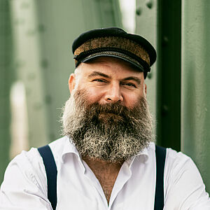 Portraitfoto von Ulrich Burdack. Er trägt ein weißes Hemd, dunkle Hosenträger, eine Schiffermütze sowie einen grauen Vollbart. Seine Arme sind vor der Brust verschränkt.