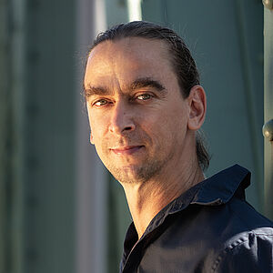 Portraitfoto von Karsten Zinser. Er steht seitlich. Sein braunes Haar ist zu einem tiefen Zopf gebunden. Er trägt ein schwarzes Hemd. Hinter ihm sind grüne Stahlstreben. 