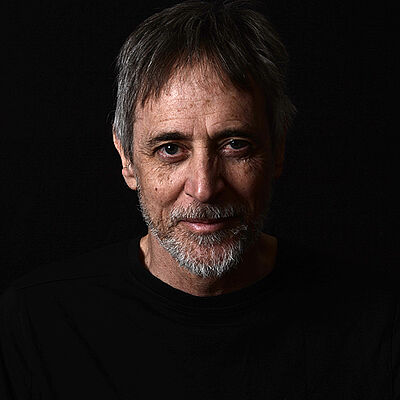 Portraitfoto von Johannes Simons. Sein graues kurzes Haar fällt ihm auf die Stirn. Er trägt einen Vollbart. Er ist in ein schwarzes Oberteil gekleidet, das mit dem schwarzen Hintergrund verschmilzt.