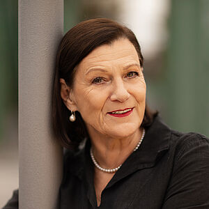 Portraitfoto von Isabel Zeumer. Sie lehnt an einer grauen Stange. Die Arme hat sie herum gelegt. Sie lächelt mit roten Lippen. Ihr braunes Haar ist schulterlang und hinter die Ohren gelegt. Sie trägt Perlenohrringe und eine Perlenkette zu einer schwarzen Bluse. 