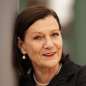Portraitfoto von Isabel Zeumer. Sie lehnt an einer grauen Stange. Die Arme hat sie herum gelegt. Sie lächelt mit roten Lippen. Ihr braunes Haar ist schulterlang und hinter die Ohren gelegt. Sie trägt Perlenohrringe und eine Perlenkette zu einer schwarzen Bluse.