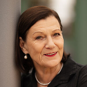 Portraitfoto von Isabel Zeumer. Sie lehnt an einer grauen Stange. Die Arme hat sie herum gelegt. Sie lächelt mit roten Lippen. Ihr braunes Haar ist schulterlang und hinter die Ohren gelegt. Sie trägt Perlenohrringe und eine Perlenkette zu einer schwarzen Bluse. 