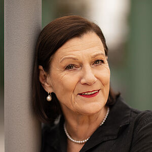 Portraitfoto von Isabel Zeumer. Sie lehnt an einer grauen Stange. Die Arme hat sie herum gelegt. Sie lächelt mit roten Lippen. Ihr braunes Haar ist schulterlang und hinter die Ohren gelegt. Sie trägt Perlenohrringe und eine Perlenkette zu einer schwarzen Bluse.