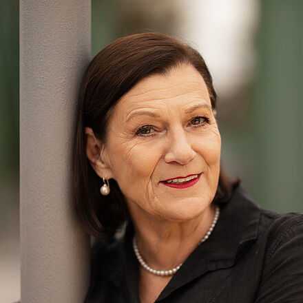 Portraitfoto von Isabel Zeumer. Sie lehnt an einer grauen Stange. Die Arme hat sie herum gelegt. Sie lächelt mit roten Lippen. Ihr braunes Haar ist schulterlang und hinter die Ohren gelegt. Sie trägt Perlenohrringe und eine Perlenkette zu einer schwarzen Bluse.