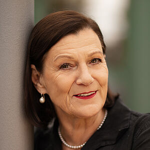 Portraitfoto von Isabel Zeumer. Sie lehnt an einer grauen Stange. Die Arme hat sie herum gelegt. Sie lächelt mit roten Lippen. Ihr braunes Haar ist schulterlang und hinter die Ohren gelegt. Sie trägt Perlenohrringe und eine Perlenkette zu einer schwarzen Bluse.