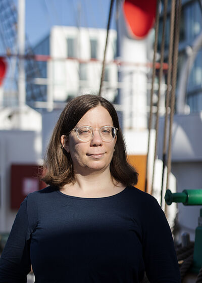 Portraitfoto von Veronika Weiser. Sie hat braunes Haar, trägt eine Brille und ein dunkelblaues Oberteil. Sie wird von der Sonne angestrahlt.