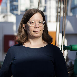 Portraitfoto von Veronika Weiser. Sie hat braunes Haar, trägt eine Brille und ein dunkelblaues Oberteil. Sie wird von der Sonne angestrahlt.