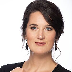 Portraitfoto von Eva Maria Summerer. Sie hat dunkelbraunes Haar, das am Hinterkopf hochgesteckt ist. Sie trägt ein schwarzes enges Oberteil mit einem V-Ausschnitt. An den Ohren baumeln tränenförmige Ohrringe. Ihre rechte Hand liegt auf dem Dekolletee. Daran trägt sie einen Ring mit schwarzem Diamanten. Der Hintergrund ist weiß.