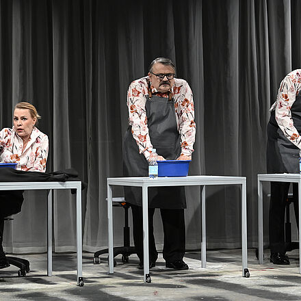 Drei Tische mit jeweils einer Person. Die Personen rechts und in der Mitte beugen sich stehend über eine blaue Schale und kneten einen Teig. Die Person links sitzt und schaut nach vorne.