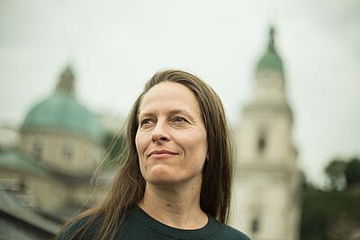 Portraitfoto von Tanja Hofmann. Sie steht vor einem Kirchengebäude, das im Hintergrund verschwommen zu erkennen ist. Sie ist von schräg unten fotografiert. Sie blickt zur Seite. Sie hat braunes langes Haar und trägt einen dunkelgrünen Pullover.