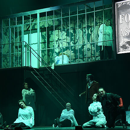 «Foggs Asylum» steht auf einer Tafel. Davor sitzen Menschen in weißen Kittel