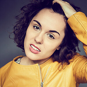 Portraitfoto von CLEO. Sie trägt eine gelbe Lederjacke und ihr lockiges kinnlanges Haar offen. Ihr Kopf ist zur Seite geneigt. Mit der linken Hand fährt sie sich durch ihr Haar. Der Mund ist leicht geöffnet. Der Hintergrund ist dunkelgrau.