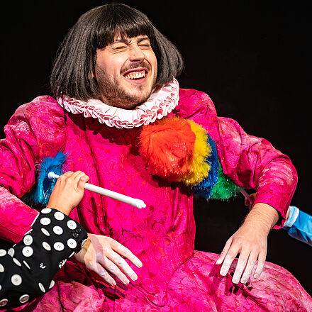 Die Person im pinken Kleid wird mit zwei Staubwedel gekitzelt.  