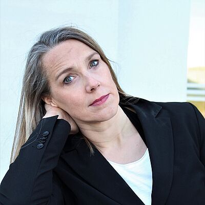 Portraitfoto einer Frau mit blonden langen Haaren. Sie trägt ein weißes T-Shirt und einen schwarzen Blazer. Sie stützt  ihren Kopf auf den rechten Arm und schaut ernst.