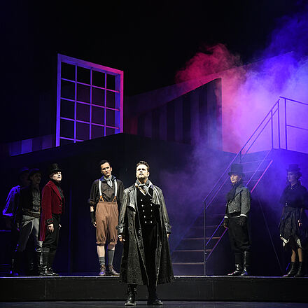 Im Hintergrund stehen mehrere Personen. Alle schauen auf den im Vordergrund stehenden Protagonisten.