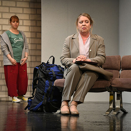 Marie Petersen steht im Hintergrund und schaut zu ihrer Mutter die vorn auf einer Sitzbank sitzt. Die Mutter, Valerie Petersen, wendet den Blick enttäuscht von ihrer Tochter ab. Marie trägt eine rote Hose, gelbe Chucks und einen grauen Hoodie. Die Mutter trägt einen beigen Hosenanzug.