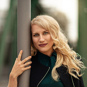 Portraitfoto von Victoria Kunze. Sie hat den rechten Arm um eine graue Säule gelegt, ihr Kopf lehnt daran. Sie trägt eine dunkelgrüne Bluse, darüber eine schwarze Jacke. Ihre blonden Wellen legen sich auf ihr Schultern.