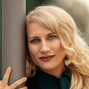 Portraitfoto von Victoria Kunze. Sie hat den rechten Arm um eine graue Säule gelegt, ihr Kopf lehnt daran. Sie trägt eine dunkelgrüne Bluse, darüber eine schwarze Jacke. Ihre blonden Wellen legen sich auf ihr Schultern.
