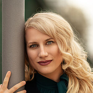 Portraitfoto von Victoria Kunze. Sie hat den rechten Arm um eine graue Säule gelegt, ihr Kopf lehnt daran. Sie trägt eine dunkelgrüne Bluse, darüber eine schwarze Jacke. Ihre blonden Wellen legen sich auf ihr Schultern.  