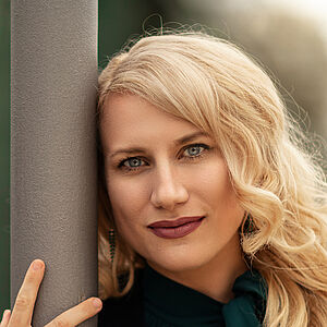 Portraitfoto von Victoria Kunze. Sie hat den rechten Arm um eine graue Säule gelegt, ihr Kopf lehnt daran. Sie trägt eine dunkelgrüne Bluse, darüber eine schwarze Jacke. Ihre blonden Wellen legen sich auf ihr Schultern. 