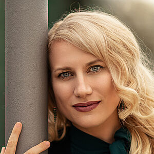 Portraitfoto von Victoria Kunze. Sie hat den rechten Arm um eine graue Säule gelegt, ihr Kopf lehnt daran. Sie trägt eine dunkelgrüne Bluse, darüber eine schwarze Jacke. Ihre blonden Wellen legen sich auf ihr Schultern.  
