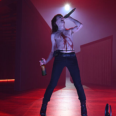 Im Hintergrund sind das Bühnenbild und rotes Licht zu sehen. Im Vordergrund steht ein singender Mann mit einem Mikrofon und einer Whiskyflasche in den Händen. Der Mann trägt kein Oberteil und hat eine rote Flüssigkeit auf der Brust.