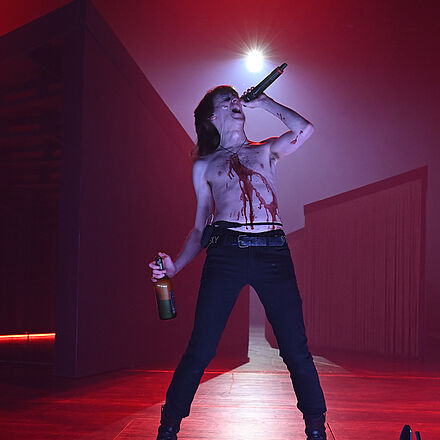 Im Hintergrund sind das Bühnenbild und rotes Licht zu sehen. Im Vordergrund steht ein singender Mann mit einem Mikrofon und einer Whiskyflasche in den Händen. Der Mann trägt kein Oberteil und hat eine rote Flüssigkeit auf der Brust.