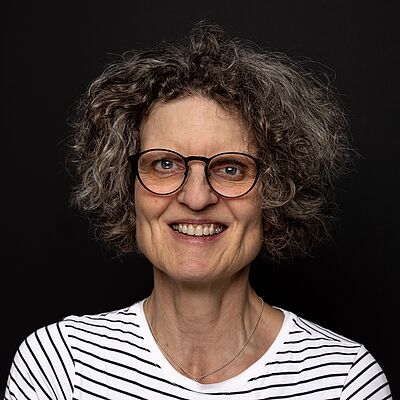 Portraitfoto von Monika Frenz. Sie hat kurze lockige Haare und trägt eine Brille sowie ein weißes T-Shirt mit schwarzen Streifen. Der Hintergrund des Fotos ist dunkel.