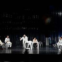 Gerichtssaal. Dorfrichter Adam und Schreiber Licht befinden sich im Zentrum. Rupert, Veit Tümpel und Gerichtsrat Walter sitzen links. Marthe Rulle und Eve sitzen rechts. Alle tragen weiße Kleidung und sitzen auf weißen Möbeln. Die Bühne ist dunkel. Dorfrichter Adam hat sich mit erhobenem Zeigefinger erhoben.