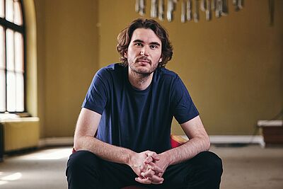 Portraitfoto von Jakob Arnold. Er sitzt auf einem Podest in einem Saal. Seine Ellbogen sind auf seine Knie gestützt. Er trägt ein dunkelblaues T-Shirt und eine schwarze Hose. Er hat kinnlanges dunkelbraunes Haar und trägt einen Bart.
