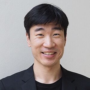 Portraitfoto von YoungMook Hwang. Er lächelt mit geöffnetem Mund. Er hält einen Dirigierstab in der rechten Hand, welchen er vor seinem Bauch hält. Er ist in einen schwarzen Anzug und schwarzes Oberteil gekleidet. Sein dunkles Haar fällt ihm auf die Stirn.