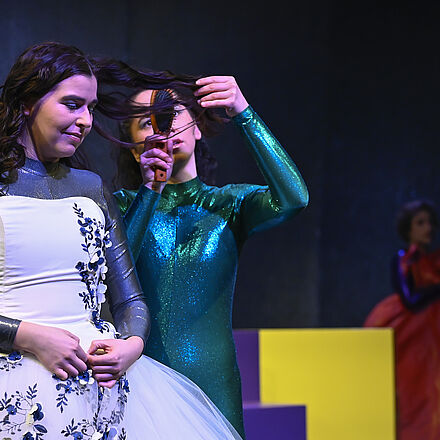 Eine Person trägt ein Prinzessinnenkleid. Ihr werden die Haare gekämmt. Im Hintergrund erkennt man die Treppen und eine weitere Person im Kleid.