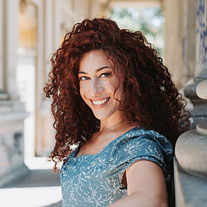 Portraitfoto von Julia Mintzer. Sie lehnt an der Wand eines marmornen Säulengangs und grinst mit offenem Mund. Ihr rotes lockiges Haar fällt ihr locker auf die Schultern. Sie steht seitlich und blickt in die Kamera. Sie trägt ein hellblaues Kleid mit weißen Aufdrucken. Durch die Säulen fällt Sonnenlicht.