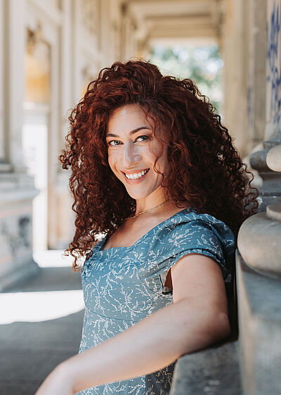 Portraitfoto von Julia Mintzer. Sie lehnt an der Wand eines marmornen Säulengangs und grinst mit offenem Mund. Ihr rotes lockiges Haar fällt ihr locker auf die Schultern. Sie steht seitlich und blickt in die Kamera. Sie trägt ein hellblaues Kleid mit weißen Aufdrucken. Durch die Säulen fällt Sonnenlicht.