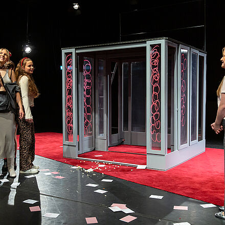 Die Mädchen stehen im Kreis Rücken an Rücken. Vor ihnen ein weiteres Mädchen. Auf dem Boden Popcorn und Karten.