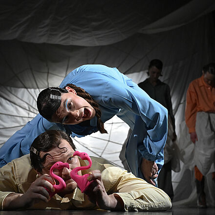 Ein Mann spielt mit Flamingos. Über ihm steht eine Frau. Im Hintergrund stehen zwei Personen, sie schauen zu Boden.
