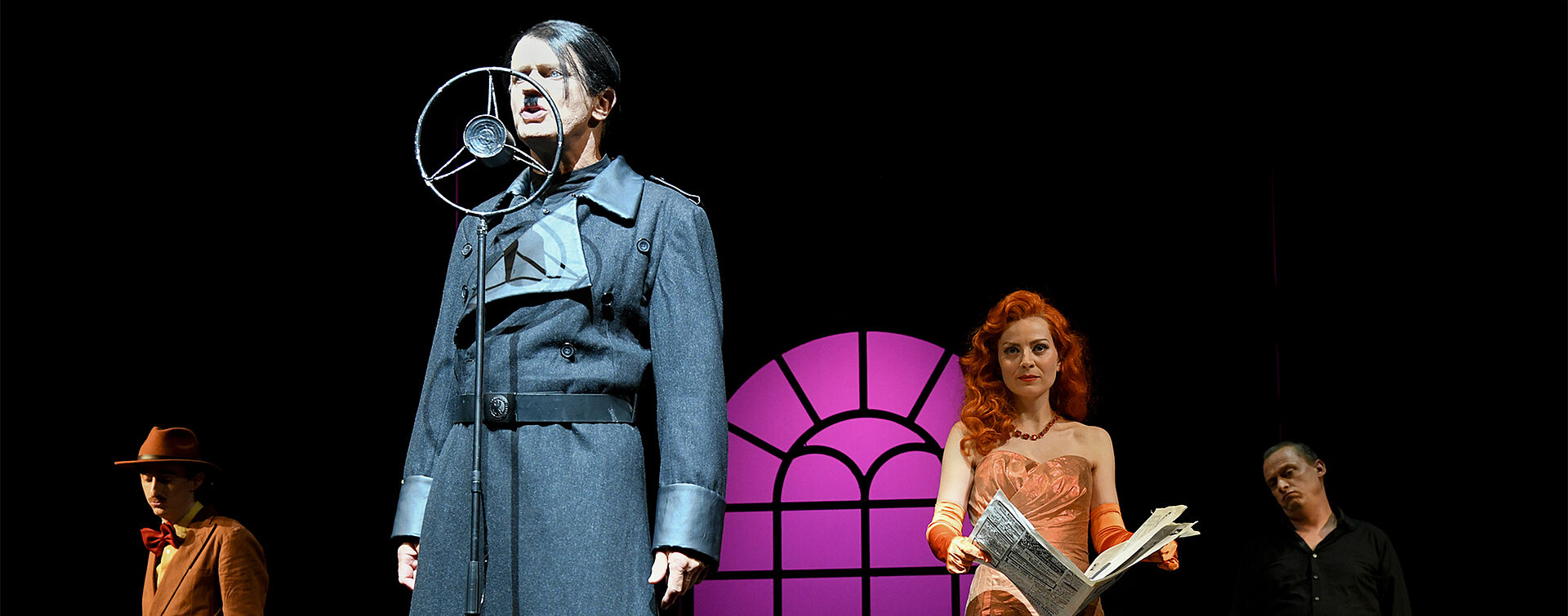 Ein bärtiger Mann mit Seitenscheitel und schwarzem Mantel steht am Mikrofon und singt. Im Hintergrund sind eine Frau in orangefarbener Kleidung, ein weiterer Mann im Mantel und ein Herr mit Hut zu sehen