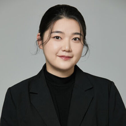 Portraitfoto von Bobin Kim. Sie ist schwarz gekleidet und trägt einen Pullover mit hohem Kragen, darüber einen Blazer. Ihr dunkles Haar ist zurückgebunden. Sie steht vor einem hellgrauen Hintergrund.