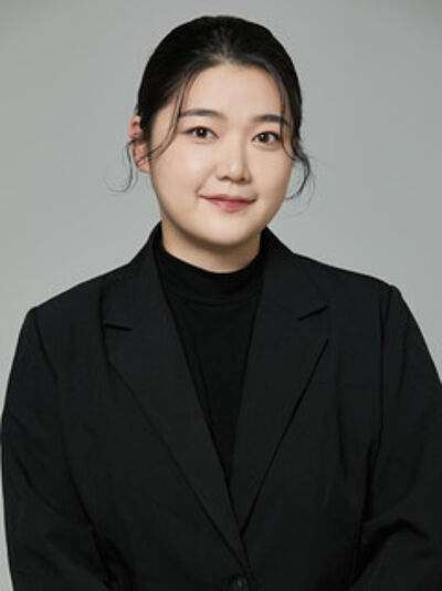 Portraitfoto von Bobin Kim. Sie ist schwarz gekleidet und trägt einen Pullover mit hohem Kragen, darüber einen Blazer. Ihr dunkles Haar ist zurückgebunden. Sie steht vor einem hellgrauen Hintergrund.