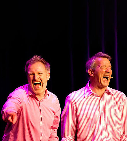 Foto zweier Mitglieder des Kollektivs "Instant Impro". Sie stehen auf der Bühne und lachen mit weit geöffnetem Mund. 