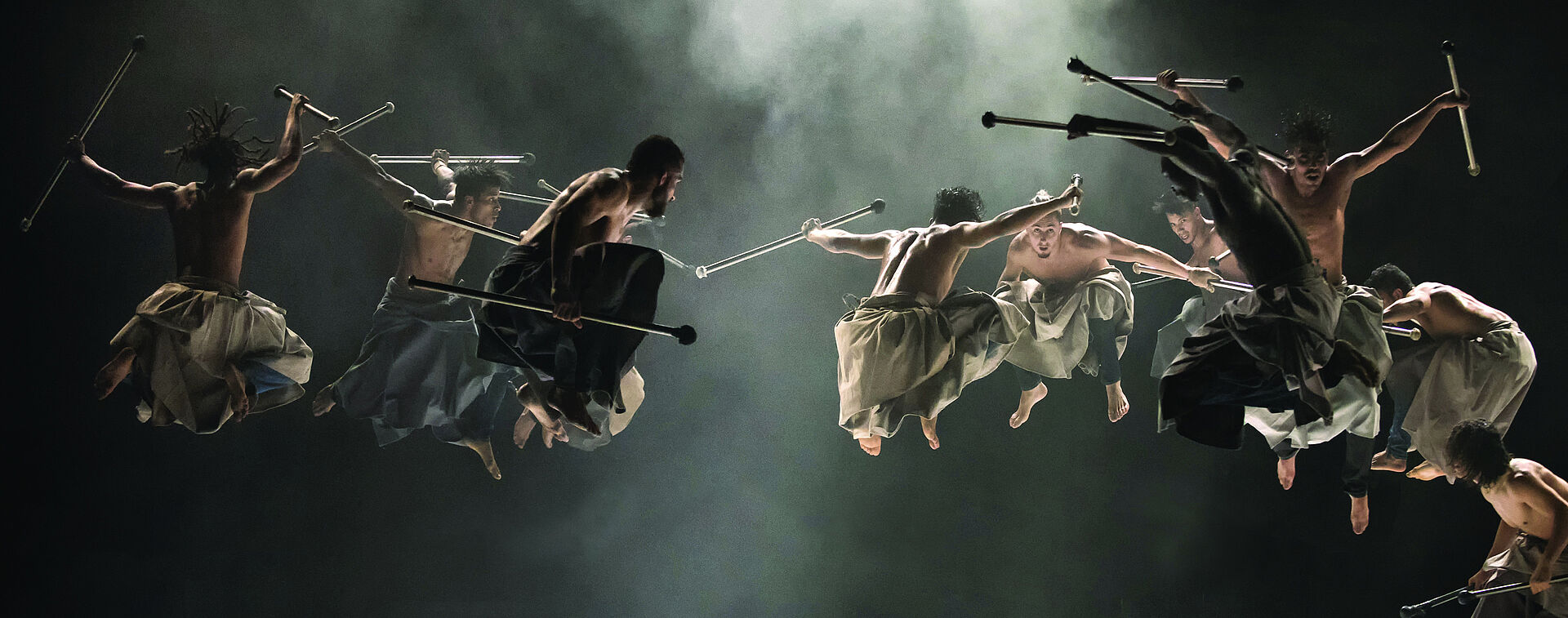 Die Tanzcompagnie "Les Nuits Barbares" im Sprung. Sie sind oberkörperfrei und haben die Knie in der Luft angezogen. In den Händen halten Sie Holzstöcke.. Sie tragen Pluderhosen. Die Bühne ist schwarz und vernebelt.