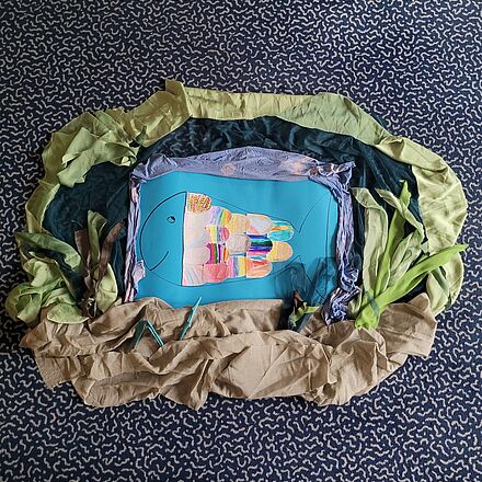 Ein gebastelter Fisch. Er hat bunte Schuppen, lächelt und außenrum wurde eine Unterwasserlandschaft gestaltet.