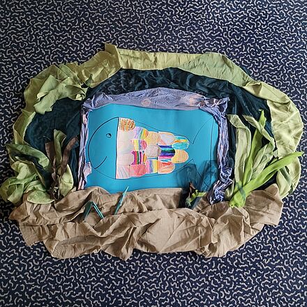 Ein gebastelter Fisch. Er hat bunte Schuppen, lächelt und außenrum wurde eine Unterwasserlandschaft gestaltet.