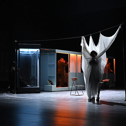 Im Haus küssen sich Menschen. Im Vordergrund steht eine Frau, gehüllt in weiße Wäsche.