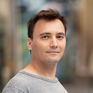 Portraitfoto von Jens Bache. Er steht seitlich und hat den Kopf nach vorn gedreht. Er hat braunes kurzes Haar und trägt ein hellgraues geripptes T-Shirt. Der Hintergrund ist verschwommen.